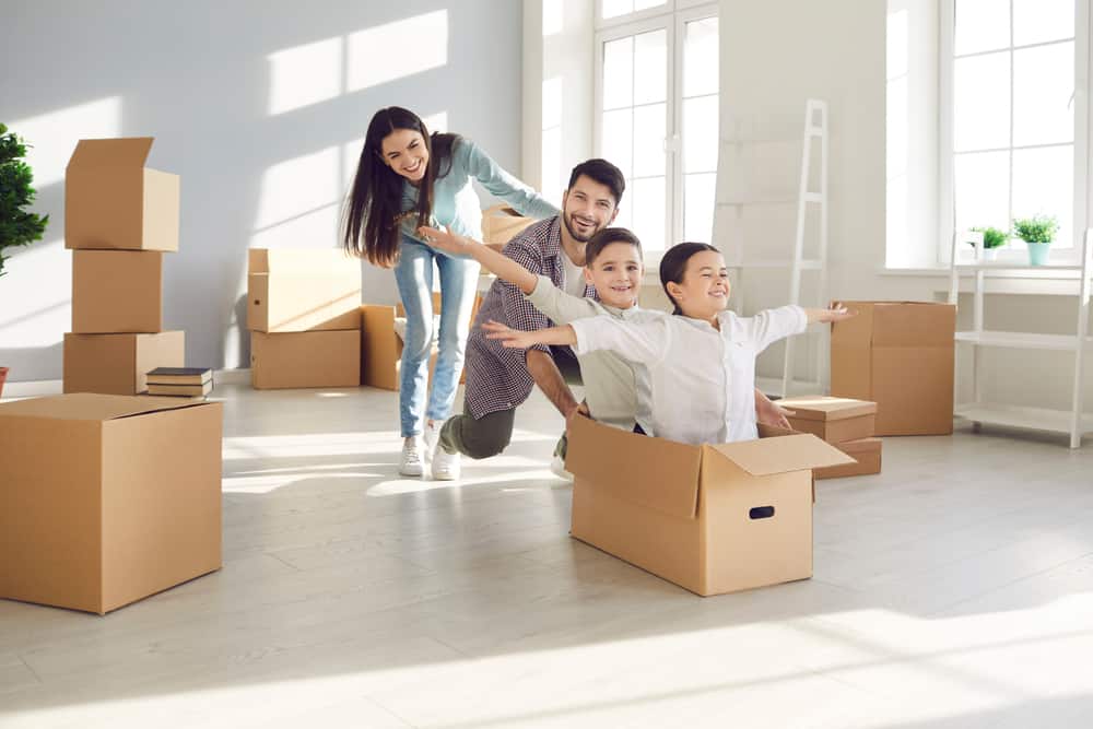A joyful family of four plays with cardboard boxes in a sunlit room. The father and mother smile as they push the two children, who are sitting inside an open box. Other packed boxes from Long Island movers are scattered around the room, suggesting they are moving or unpacking.