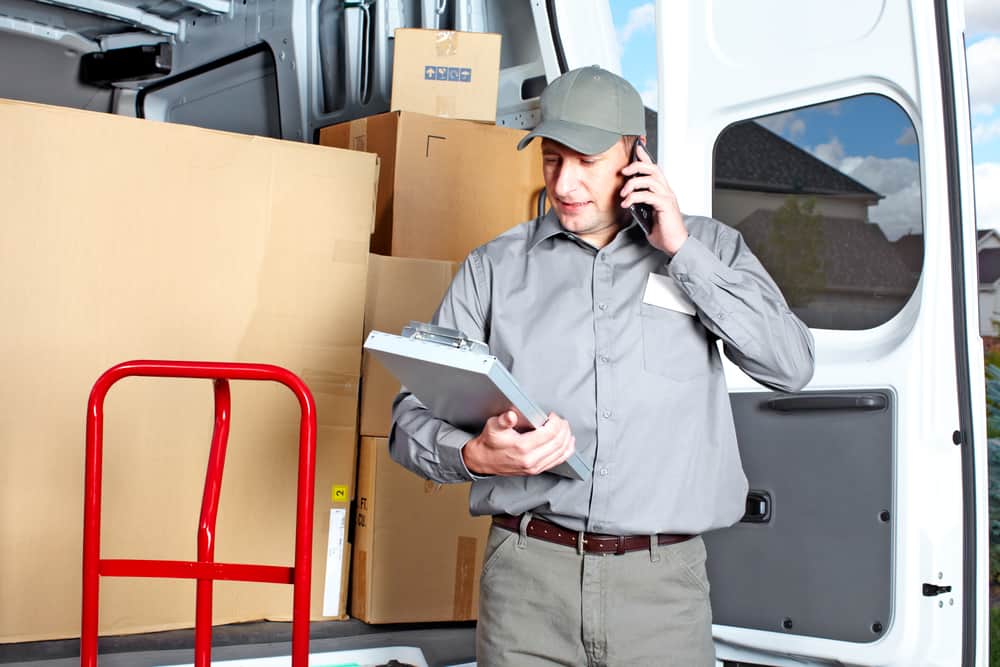 A delivery person wearing a gray uniform and cap is holding a clipboard and talking on a mobile phone beside an open delivery van. The van, typical of those used by Long Island moving companies, is filled with cardboard boxes, and a red hand truck is positioned nearby. The sky and a house are visible in the background.