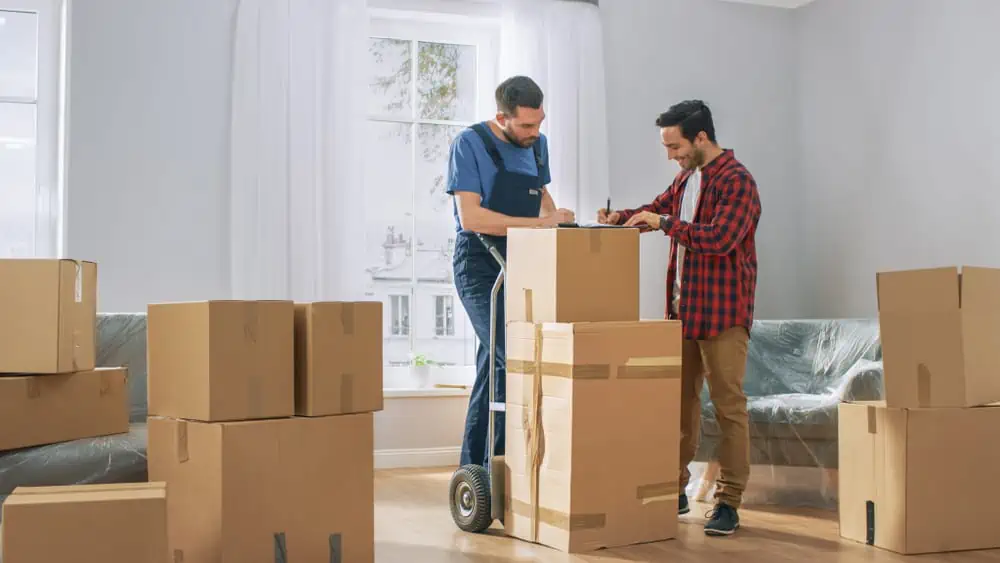 Two men are standing in a room filled with moving boxes. One man is holding a clipboard while the other signs a document. They appear to be utilizing Long Island movers for their move or delivery. The room has a large window with light curtains and a plastic-covered sofa.