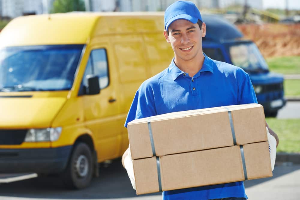 A delivery person wearing a blue uniform and cap smiles while holding two cardboard boxes. Behind him, there are yellow and blue delivery vans parked on the sunny street. It's evident that Long Island moving companies take pride in their efficient, friendly service.