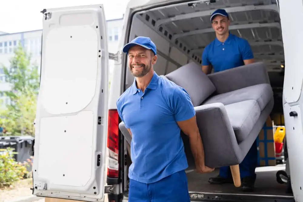 Two movers in blue uniforms, working for a Long Island moving company, are carrying a gray sofa from the back of a white moving van. Both are smiling as they work, with one at each end of the sofa. The back doors of the van are open, and a building is visible in the background.