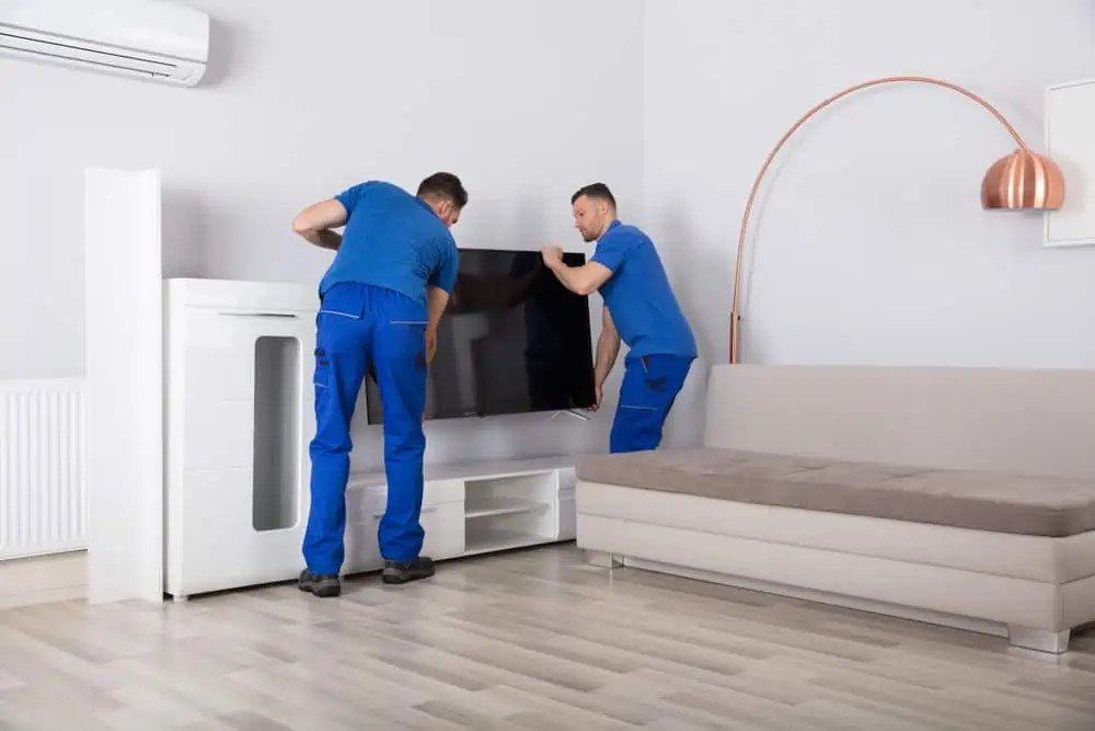 Two workers in blue uniforms, from a reputable Long Island moving company, are installing a flat-screen TV onto a white TV stand in a modern living room. The room features a light-colored sofa, a floor lamp with a copper finish, and a wall-mounted air conditioner.