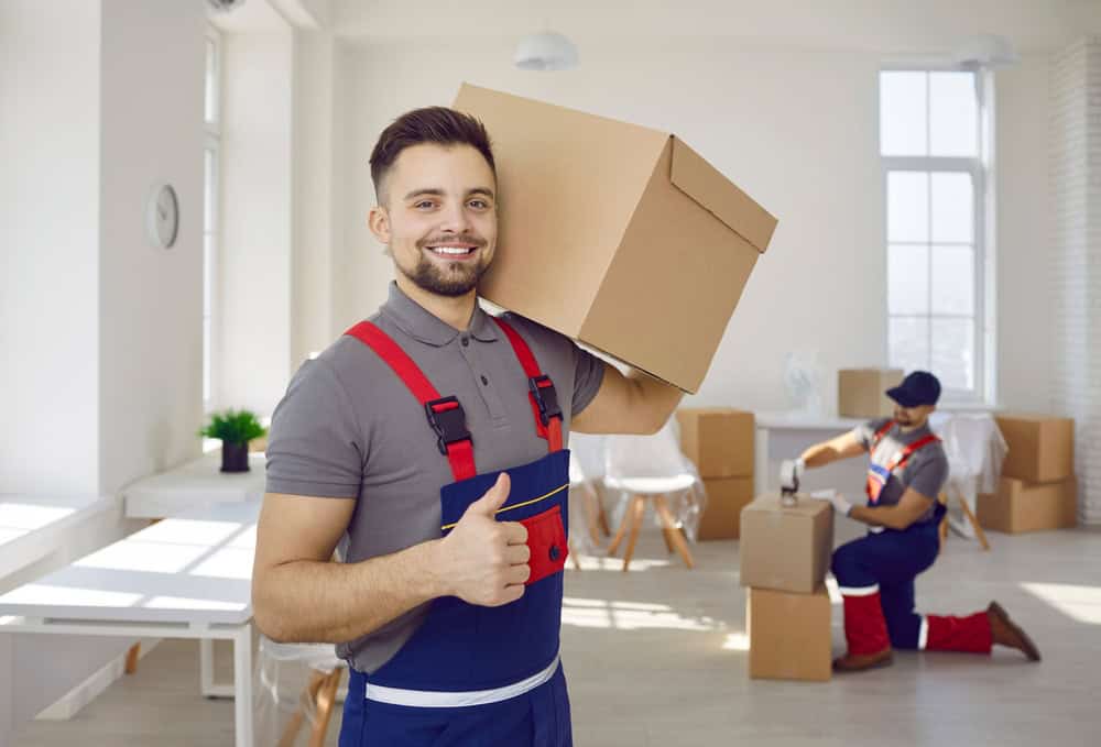 A man in a gray shirt and red suspenders smiles and gives a thumbs-up while carrying a cardboard box on his shoulder in a well-lit room. Another man in similar attire, from Long Island moving companies, is taping boxes in the background.