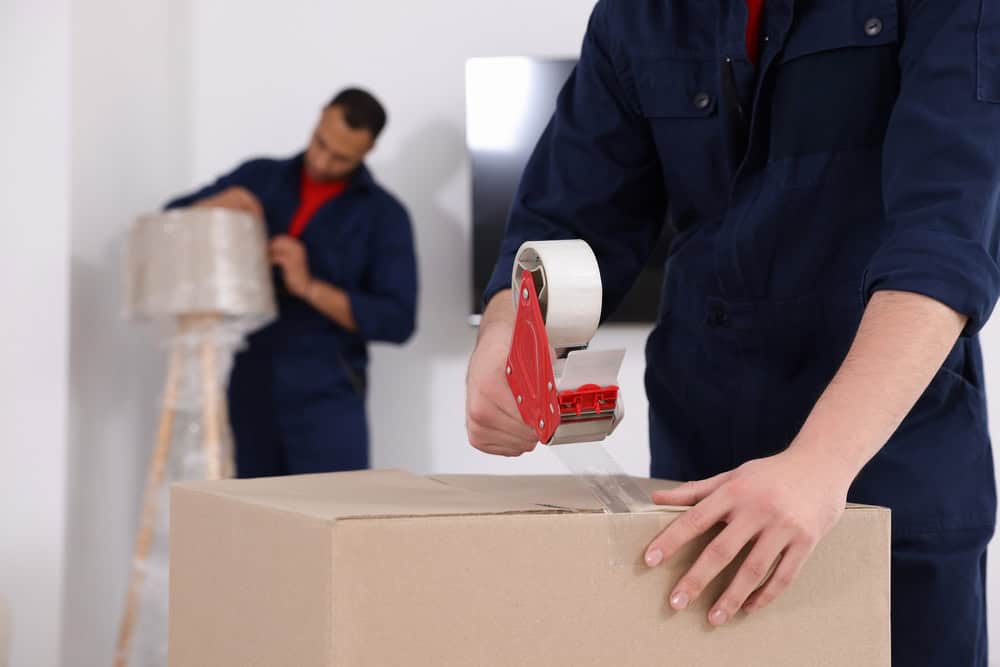 Two people working in a room. One in the foreground is sealing a cardboard box with packing tape. The other in the background is wrapping a lamp with protective bubble wrap. Both, dressed in dark blue uniforms, are part of a dedicated team from Long Island movers ensuring items are secure for transport.