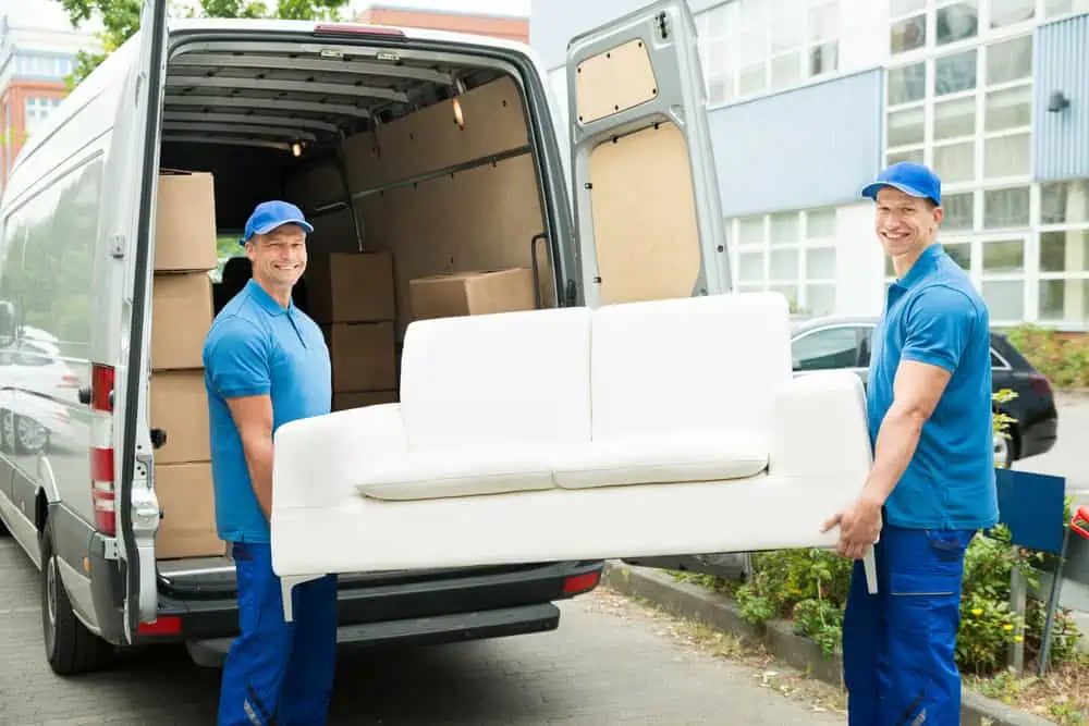 Two movers from a Long Island moving company, wearing blue uniforms and caps, are smiling while carrying a white couch from a van filled with cardboard boxes. The van’s back doors are open, and the background shows a building with windows.